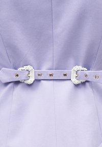 Ύφασμα λεβάντας με ταιριαστή ζώνη που διαθέτει δύο περίτεχνες λευκές αγκράφες και μεταλλικές οπές, παρουσιάζοντας λεπτομερή ραφές και υφή.