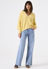 Mujer de pie con un suéter de punto grueso amarillo, jeans de pierna ancha azul claro y zapatillas grises frente a un fondo blanco liso.