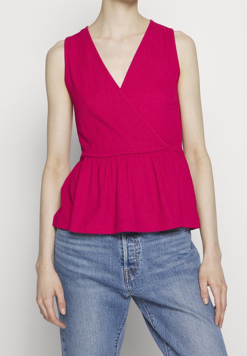 Femme portant un top sans manches rose vif à col en V avec basque, associé à un jean denim bleu clair sur un fond uni.