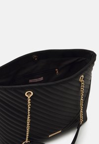 Black quilted tote bag made from synthetic material, featuring gold chain accents and a zippered inner pocket labeled "ANNA FIELD."