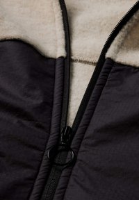 Fleece- und Polyesterjacke mit einem schwarzen Reißverschluss, einem beigen Plüschkragen und einem gesteppte dunklen Körper mit subtiler Textur.