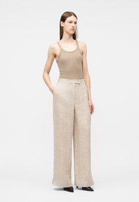 Jeune femme debout, les mains dans les poches, portant un débardeur beige, un pantalon large en lin clair et des talons noirs à bouts pointus sur fond blanc.