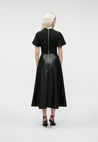 Femme aux longs cheveux blonds portant un haut noir à manches courtes et une jupe midi en cuir noir avec fermeture éclair au dos, accompagnée de sandales à talons hauts noires.