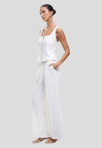 Top bianco senza maniche con zip frontale e scollatura quadrata, abbinato a pantaloni bianchi larghi con tasche laterali.