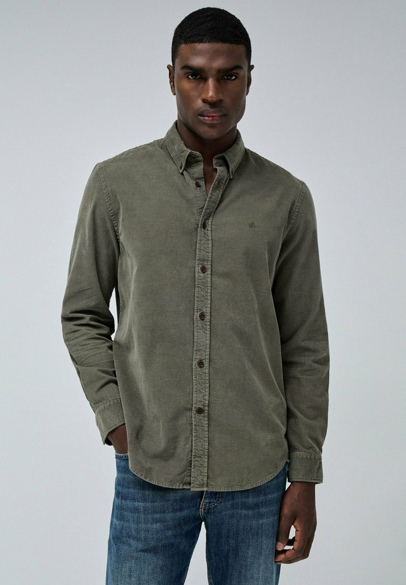 Camisa de botones verde oliva hecha de una tela suave, con un cuello clásico, botones marrones y mangas largas, combinada con pantalones vaqueros de denim azul.