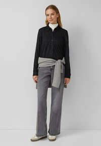 Zwarte blouse met gestructureerde textuur, grijze trui om de taille geknoopt, lichtgrijze wijde jeans en beige schoenen met witte accenten.