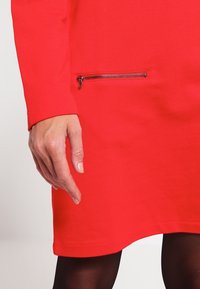 Rode jurk van gestructureerde stof, met lange mouwen en een zijzak met ritssluiting. De zoom is afgerond, met een aansluitende snit.