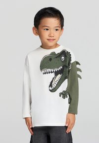 Biała koszula z długim rękawem z zielonym nadrukiem dinozaura z przodu oraz ciemnozielonymi rękawami. Materiał wydaje się miękki; luźny krój.