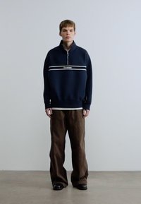 Μπλε ναυτικό φλις φούτερ με φερμουάρ ως το ένα τέταρτο, λευκές οριζόντιες ρίγες και λογότυπο στο κέντρο, συνδυασμένο με καφέ παντελόνι με φαρδιά πόδια και μαύρα παπούτσια.