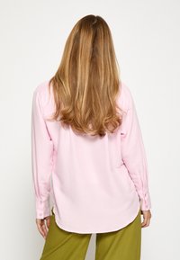 Camicia rosa chiaro a maniche lunghe con orlo rotondo e polsini abbottonati, realizzata in un tessuto morbido e texturizzato. Indossata sopra pantaloni verde oliva.