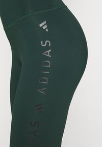 Mörkgröna Adidas leggings med vertikala svarta och reflekterande Adidas-logotyper och text på utsidan av låret.