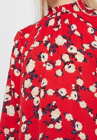 Blouse fleurie rouge avec un col montant, ornée de roses blanches et de feuilles bleues. Le tissu semble doux avec une texture lisse et des détails froncés.