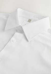 Chemise blanche texturée à col avec patte de boutonnage, soigneusement pliée, montrant le col et une partie du devant.