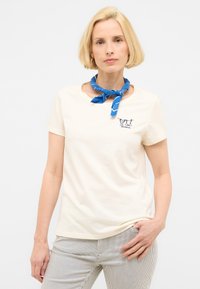 Camiseta de manga corta color beige con escote redondo y pequeño diseño bordado en azul. Combinada con pantalones de color claro a rayas.