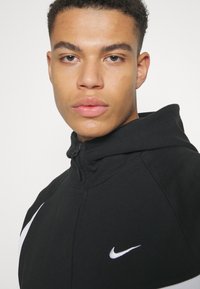 Čierna mikina Nike s vysokým golierom, na ktorej je na prednej strane biely logo swoosh. Textúrovaná tkanina so zipsom a moderným dizajnom.