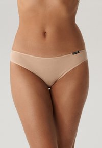 Beżowe, bezszwowe majtki bikini o niskim stanie, wykonane z miękkiego materiału, z małą czarną metką marki na pasie.