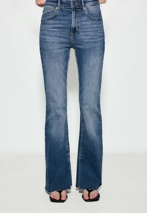Jean bleu taille haute évasé avec poches avant et ourlet brut, porté avec des sandales noires à bouts ouverts sur une personne debout.