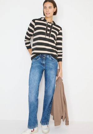 Stribet hættetrøje i sort og beige med snoretræk, kombineret med blå denimjeans og holder en brun jakke. Hvide sneakers fuldender looket.