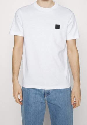 Mężczyzna w białej, gładkiej koszulce z krótkim rękawem, z czarnym kwadratowym logo na piersi i jasnoniebieskich dżinsach, stoi na tle białego tła.