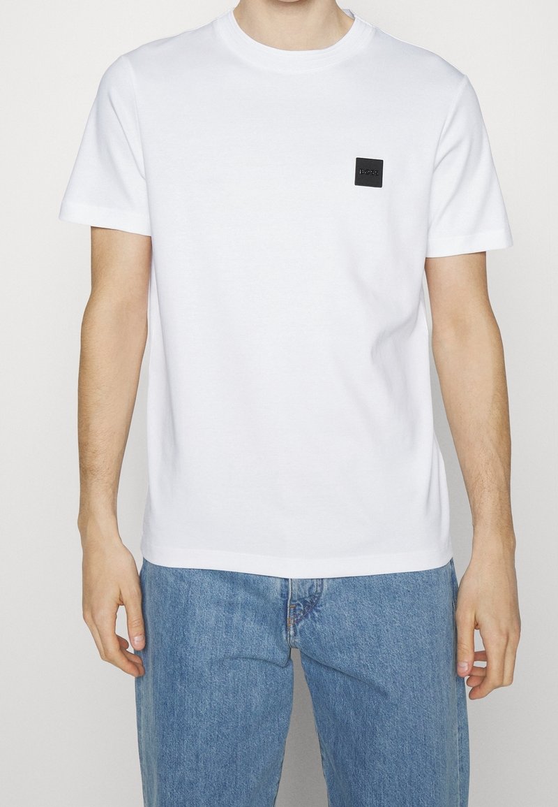 Homme portant un t-shirt blanc uni à manches courtes avec un logo carré noir sur la poitrine et un jean en denim bleu clair, debout devant un fond blanc.