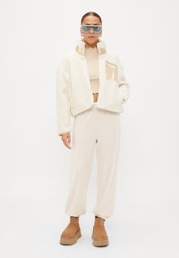 Jachetă scurtă din blană sintetică albă cu un buzunar lucios, top scurt bej, pantaloni bej și botine cu platformă maro. Ochelarii de soare gri completează look-ul.