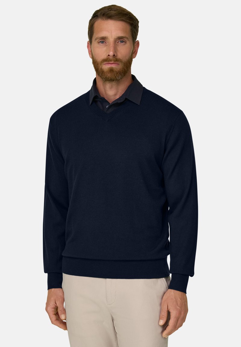 Homme aux cheveux bruns courts et à la barbe, portant un pull noir à col en V sur une chemise noire à col et un pantalon beige, debout devant un fond blanc.