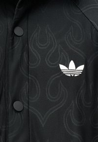 Μαύρο ύφασμα με διακριτικά σχέδια φλόγας, δύο μαύρα κουμπιά και λευκό κεντημένο λογότυπο Adidas trefoil στη δεξιά πλευρά.