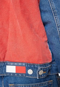Närbild av en röd och blå denimjacka som visar Tommy Jeans-märket och metalldetaljer på baklivets midja.