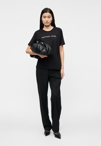 T-shirt preta em algodão com o logo "MICHAEL KORS", combinada com calças pretas de cintura alta e uma grande mala preta despojada.