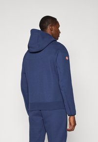 Marineblå hettegenser med ribbestrikkede mansjetter og kant, med en liten logopatch på venstre skulder og glatt tekstur.