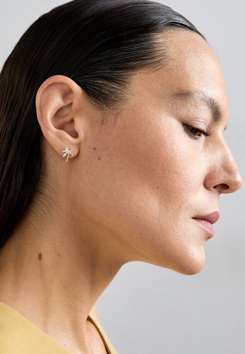 Złote kolczyki z delikatnym wzorem liścia, ozdobione małymi, przezroczystymi kryształkami. Lśniąca tekstura i skomplikowane detale.