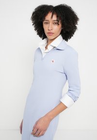 Lyseblå polo-kjole med et teksturert stoff, som har hvit krage, en liten brodert logo på brystet og lange ermer.