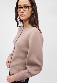 Νεαρή γυναίκα με μακριά σκούρα μαλλιά και γυαλιά, φορώντας μια μπεζ κάρντιγκ με φουσκωτά μανίκια, στέκεται μπροστά σε μία απλή ανοιχτή φόντο.