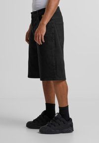 Shorts de denim negro con un corte relajado, a la altura de la rodilla, con bolsillos laterales y un acabado texturizado, combinados con zapatillas negras y calcetines.