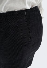 Jupe en velours côtelé noire avec une fermeture éclair sur le côté portée avec une chemise blanche rentrée sur une personne.