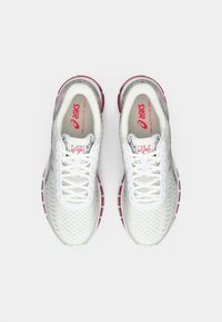 Chaussures de sport blanches avec une tige en maille respirante, rehaussées de détails rouges sur la semelle et les lacets, présentant un bout rond et un design à lacets.
