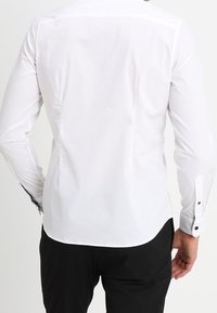 Camicia bianca con collo a bottoni, dalla texture liscia, vestibilità sartoriale e orlo arrotondato. Presenta dettagli contrastanti sui bottoni e sui polsini.