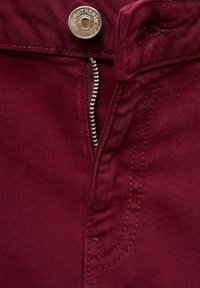 Tissu bourgogne avec une braguette zippée et une fermeture à bouton en métal, avec des coutures visibles le long des bords et sur le côté. Texture lisse.