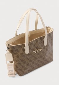 Bolso tote de lona marrón con detalles y asas de cuero beige claro, presenta un logo dorado "Guess" y una correa ajustable.