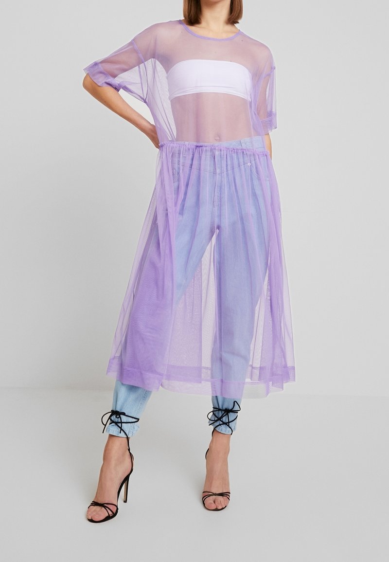 Superposition violette transparente avec des manches courtes, superposée sur un top blanc et un jean en denim bleu clair. Présente une taille froncée et des détails à lacets aux chevilles.