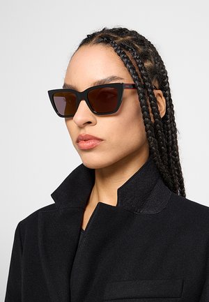 Donna con i capelli intrecciati che indossa occhiali da sole neri a occhi di gatto e un cappotto nero con collo alto, che guarda leggermente di lato.