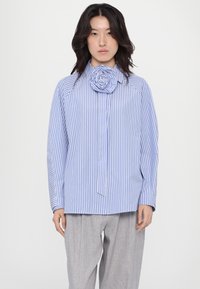 Camicia a righe blu chiaro e bianco con una grande rosa di tessuto all'collo. Maniche lunghe e vestibilità morbida, abbinata a pantaloni grigi.