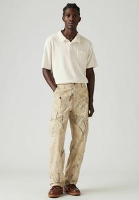 Vaaleanbeige lyhythihainen polo-paita, jossa on tasku; kuvioidut beige cargo-housut, joissa on sivutaskut; ruskeat kengät, joissa on teksturoidut pohjat.