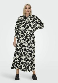 Zwart-wit gestreepte maxi jurk met lange mouwen, knoopsluiting aan de voorkant, losse pasvorm en grote zwarte vlekken op een ivoorkleurige achtergrond.