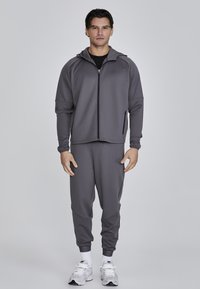 Sivá športová bunda na zips s kapucňou, zodpovedajúce sivé tepláky, vyrobené z hladkého materiálu, s jemným logom a rebrovanými manžetami.