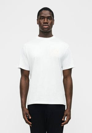 Musta mies seisoo ja katsoo eteenpäin, pukeutunut yksinkertaiseen valkoiseen lyhythihaiseen t-paitaan ja mustiin housuihin, neutraali ilme, vaalea tausta.