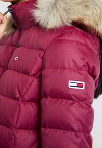Bordowa kurtka puchowa z kapturem wykończonym futrem. Posiada pikowane sekcje oraz naszywkę z logo na rękawie. Zapinana na zamek przy szyi.