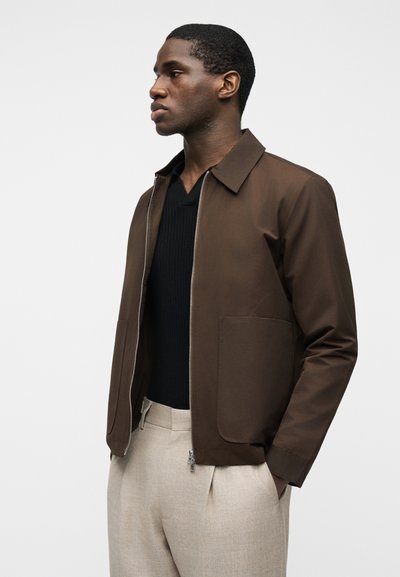 Άνδρας που φοράει καφέ ζακέτα με φερμουάρ πάνω από μαύρο πουλόβερ με V λαιμόκοψη και μπεζ παντελόνι, στέκεται με τα χέρια στις τσέπες, κοιτάζοντας προς τα αριστερά.