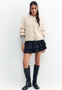 Femeie purtând un cardigan bej cu nasturi peste un top gri cu mâneci lungi, o fustă mini în carouri negre cu volane și cizme negre până la genunchi, stând în picioare.