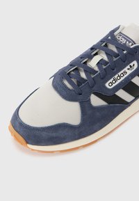 Adidași din suede albastru și plasă, cu accente albe și dungi negre, prezintă o talpă din cauciuc și închidere cu șireturi, cu un logo de marcă pe lateral.